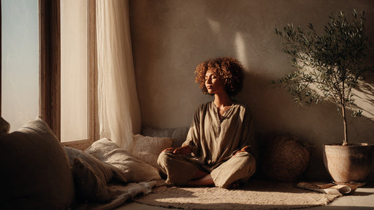 Woman meditating in a sunlit room, sitting cross-legged on a cushion with neutral pillows and a potted olive tree, creating a calm and natural atmosphere