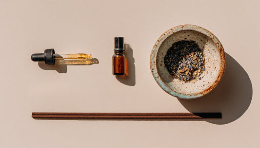 A flatlay composition showing a dropper of essential oil, a small amber glass bottle, a ceramic bowl filled with dried botanicals, and incense sticks arranged on a soft neutral surface under warm light.